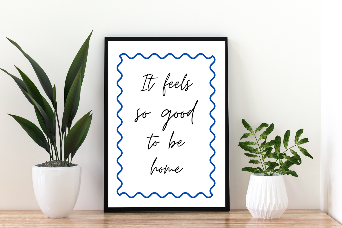 Framed print with 'It feels so good to be home' text between two potted plants on a wooden surface. Motivational quote art print with cobalt blue swirls, framed and placed in a bright, airy hallway with contemporary décor.