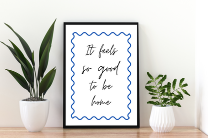 Framed print with 'It feels so good to be home' text between two potted plants on a wooden surface. Motivational quote art print with cobalt blue swirls, framed and placed in a bright, airy hallway with contemporary décor.