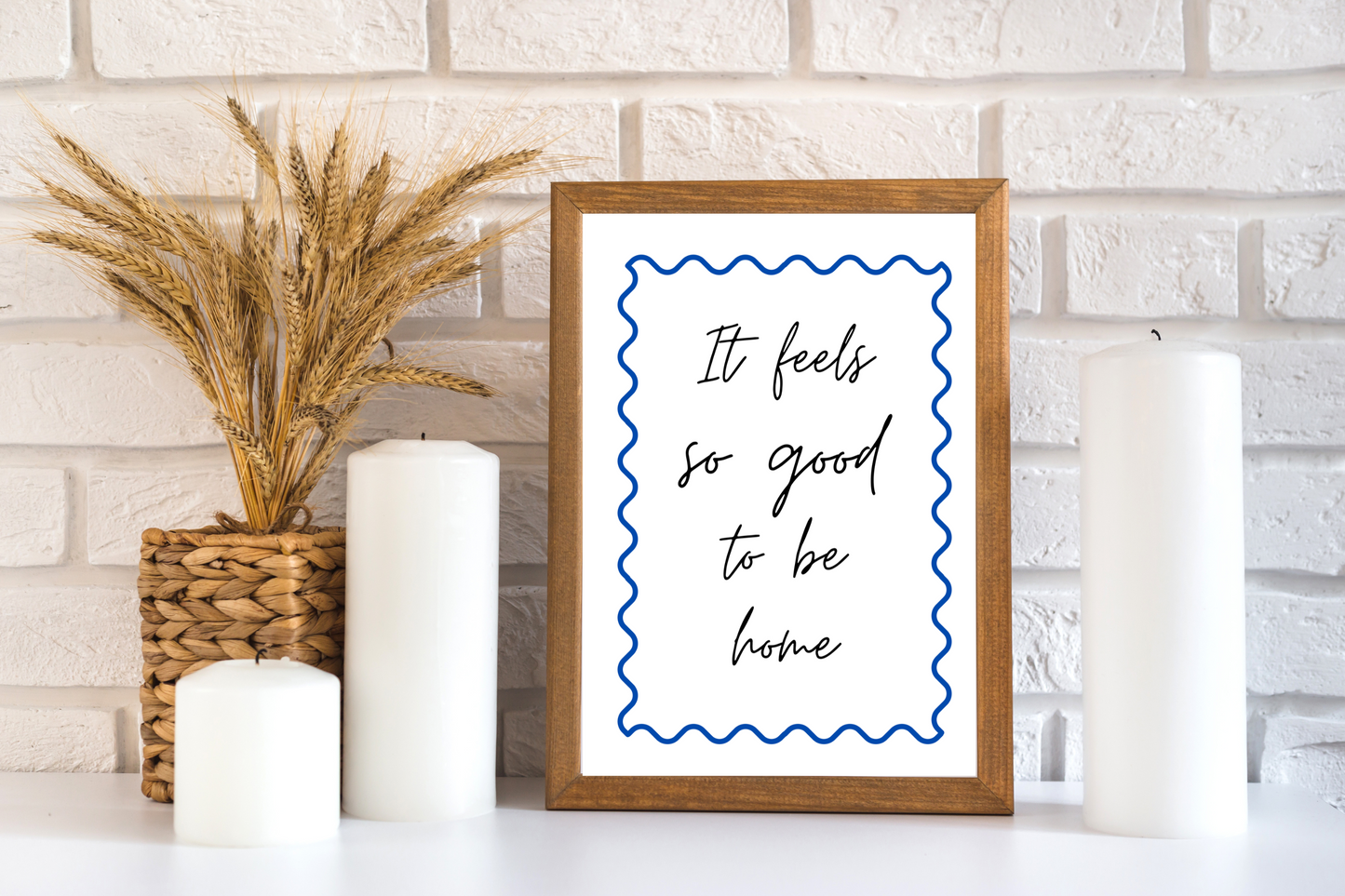 Framed print with 'It feels so good to be home' text on a white brick wall background. Printable soft life quote with blue swirly design, elegantly displayed in a boho-styled bathroom against a wall next to pillar candles and a pampas grass plant.