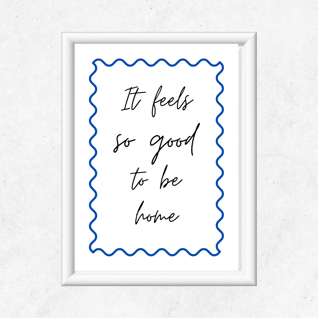 Framed print with the quote 'It feels so good to be home' on a white background with a cobalt blue swirly design. Inspirational typography print in a white frame hanging on a clean Scandinavian white wall in a feminine home office with natural light.