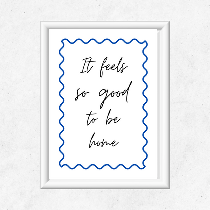 Framed print with the quote 'It feels so good to be home' on a white background with a cobalt blue swirly design. Inspirational typography print in a white frame hanging on a clean Scandinavian white wall in a feminine home office with natural light.