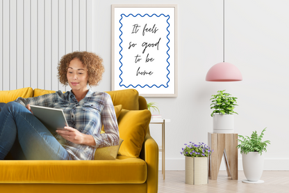 Woman sitting on a comfortable mustard yellow couch reading a tablet in a cosy living room with a motivational poster with the caption "It feels so good to be home." A printable wall art framed in a modern, minimalist reading corner with plant-filled surroundings, perfect for a soft life aesthetic.